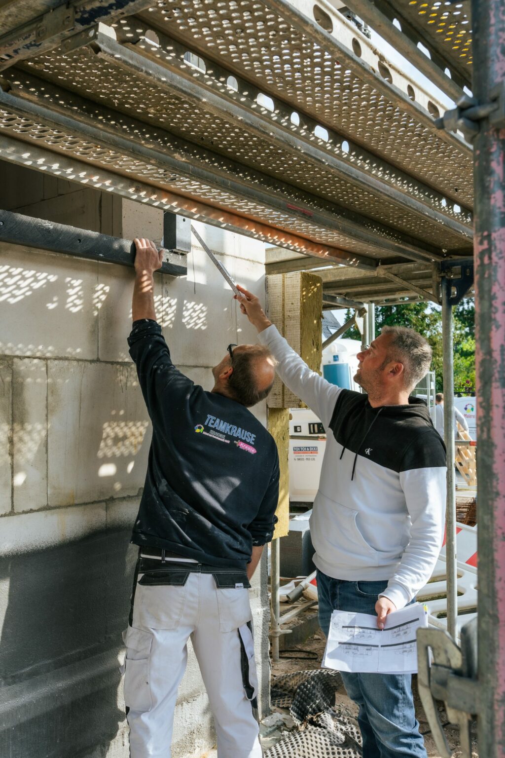 Zwei Mitarbeiter von Malermeister Krause überprüfen auf einer Baustelle die Wandisolierung unter einem Gerüst.