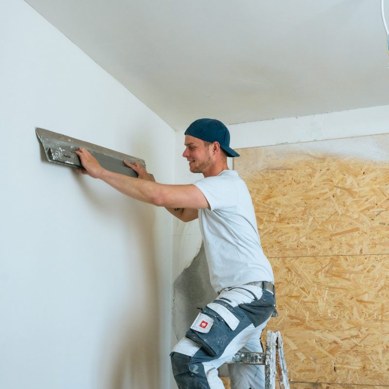 Ein junger Maler verputzt eine Wand mit einer Glättkelle und lächelt dabei.